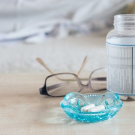 A bottle of supplement tablets next to a small dish with two white pills and a pair of glasses on a nightstand.