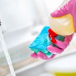 Someone using dish gloves, squeezing dish soap onto a sponge besides a sink with running water.