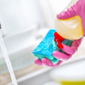 Someone using dish gloves, squeezing dishwashing detergent onto a sponge besides a sink with running water.