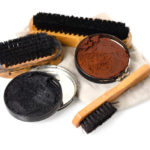 A tin of both black and brown shoe polish sits on a shining cloth besides polishing brushes.