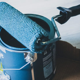 A roller sitting on top of an open bucket of wall paint.