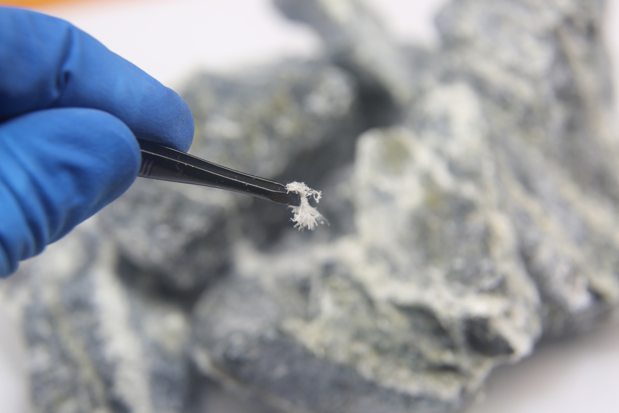 Someone wearing gloves holds a piece of asbestos with tweezers.