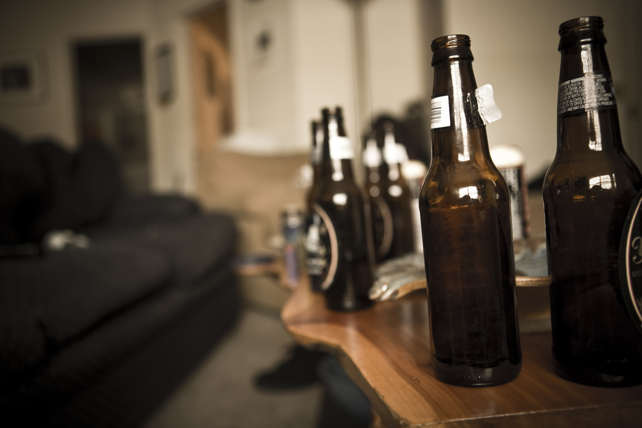 A living room with a table in front of a couch full of empty beer bottles on it with someone blurred in the background experiencing alcohol poisoning.
