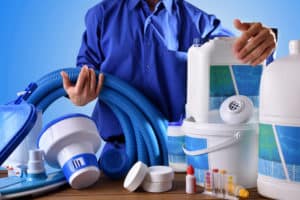 Person in a blue shirt handling large containers of chlorine-based pool chemicals, surrounded by tablets, granules, hoses, and water testing supplies on a table near a pool.
