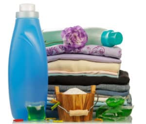 An assortment of laundry products including a blue liquid detergent bottle, stacked folded towels, a wooden bucket, fabric softener, and dryer sheets arranged together.
