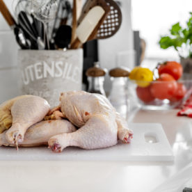 Raw chicken thighs sit on a cutting board on a counter besides tomatoes to represent how uncooked meat can become spoiled food.