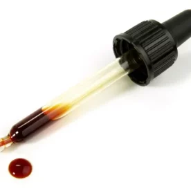 A glass dropper holds a bit of iodine at its tip. The dropper is on a white background and the iodine is brown. There's one droplet of iodine beside the dropper.