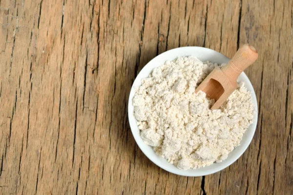 A bowl of colloidal oatmeal with a wooden scooper set on it. The bowl is resting on a wooden table.