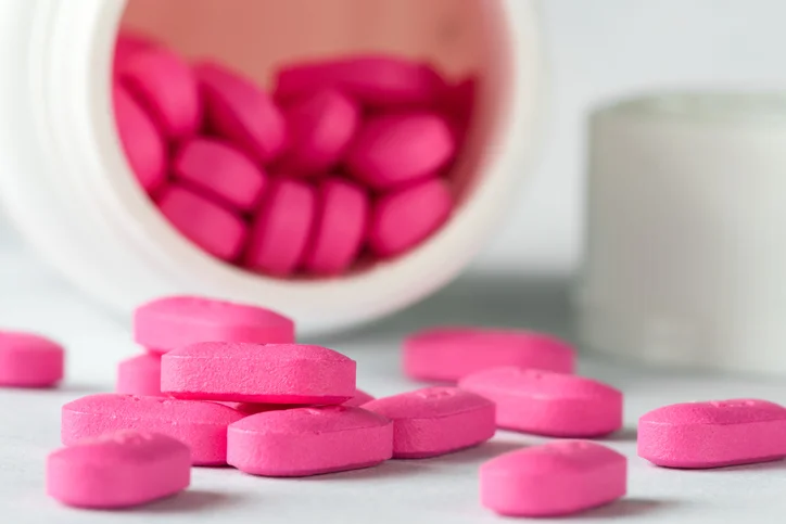 Pink pills spilled onto a white table. The pill bottle is on its side in the background. A small pile of spilled pills are in focus in the front.