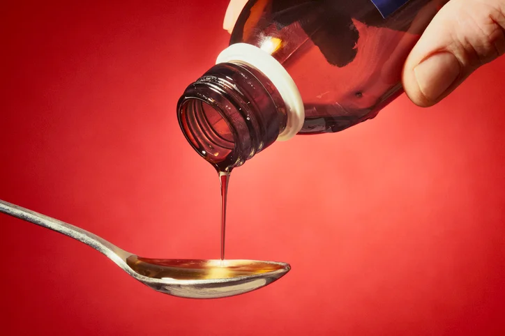 Someone pours syrup from a red bottle onto a metal spoon. The background is also red.
