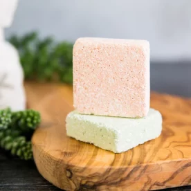 Two shower steamers arranged on a wooden board in a spa-like environment.