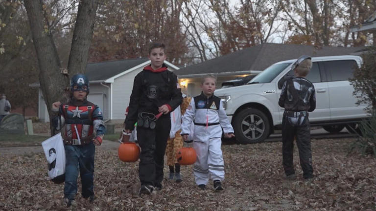Four children in Halloween costumes holding orange pumpkin buckets while trick-or-treating in a residential neighborhood at dusk
